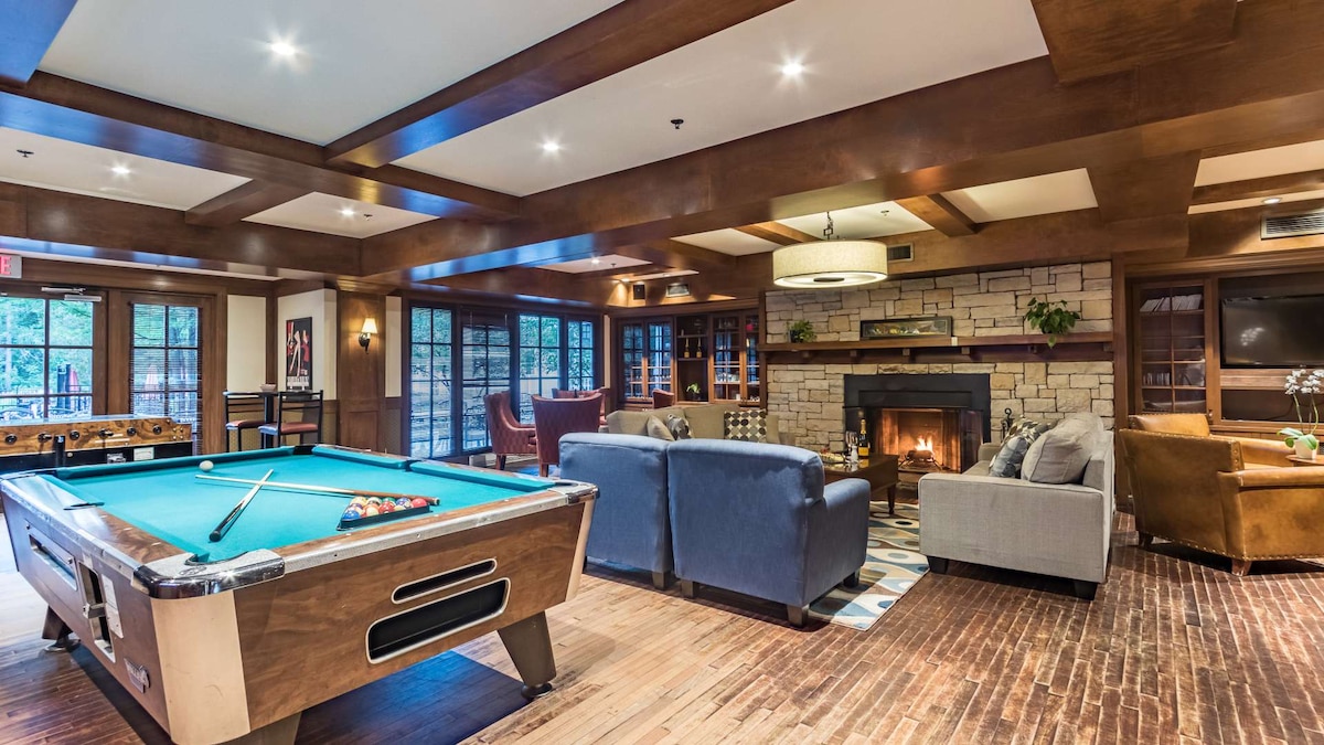 A welcoming lounge area features a classic pool table in the foreground, surrounded by comfortable seating arrangements. A warm fireplace is set within a stone wall, and large windows allow natural light to filter through, enhancing the cozy atmosphere of the space.