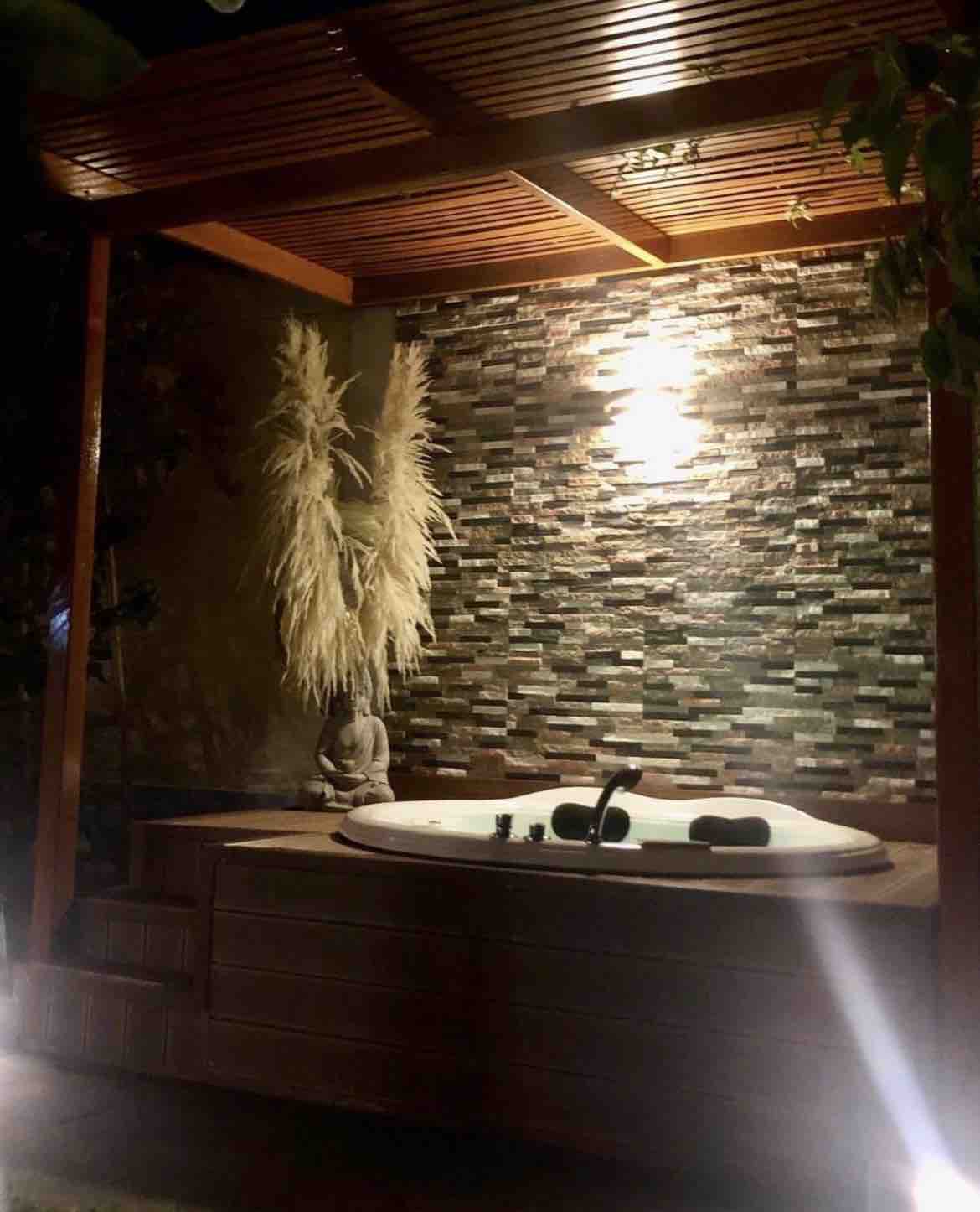 A serene outdoor space features a hot tub surrounded by natural stone walls. Soft lighting highlights decorative plants and a tranquil sculpture, creating a peaceful ambiance for relaxation.