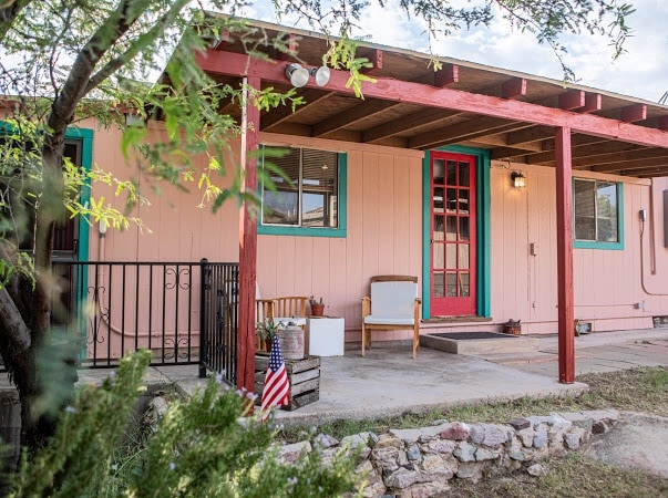 The exterior of the bunkhouse features a welcoming porch with a vibrant red door and light pink walls. A bench and small chair are situated on the patio, surrounded by greenery and a rock border. A small American flag adds a touch of character.