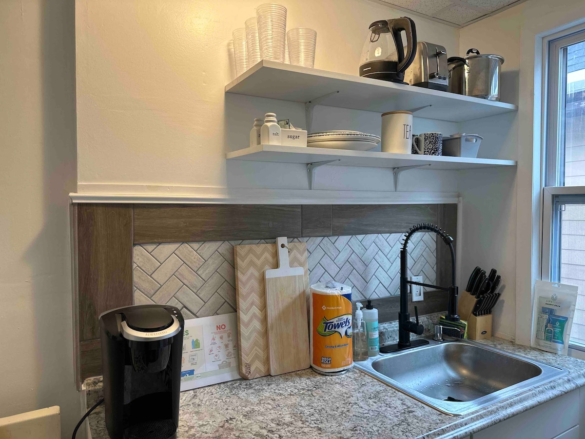 A modern kitchen space features a countertop with a stainless steel sink and neatly organized dishes. Above, white shelves hold a coffee maker, kettle, and glassware. A cozy backsplash with a herringbone design adds subtle texture. Natural light filters through the nearby window.