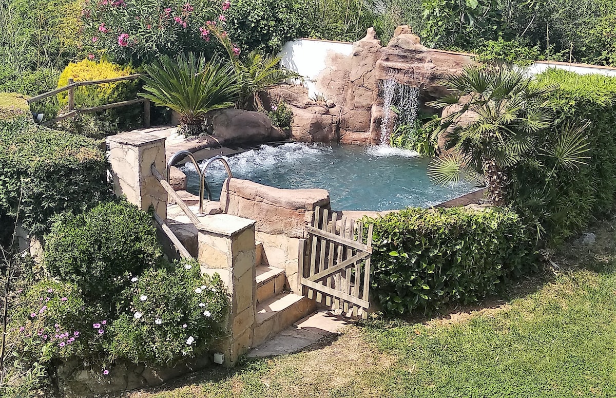 An outdoor jacuzzi is surrounded by lush greenery, tropical plants, and flowering bushes. Water cascades gently from a rock formation, creating a serene atmosphere. Steps lead down to the jacuzzi, framed by a wooden gate and natural stone. Sunlight highlights the clear water.