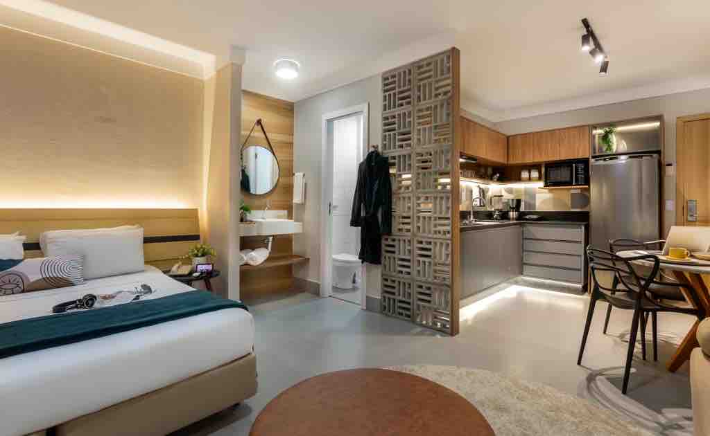 A modern studio apartment is featured, with a spacious bed positioned near an elegant wall panel. The kitchen area is equipped with contemporary appliances and sleek wooden cabinetry. A small dining table with chairs is visible, creating a functional and cozy living space.