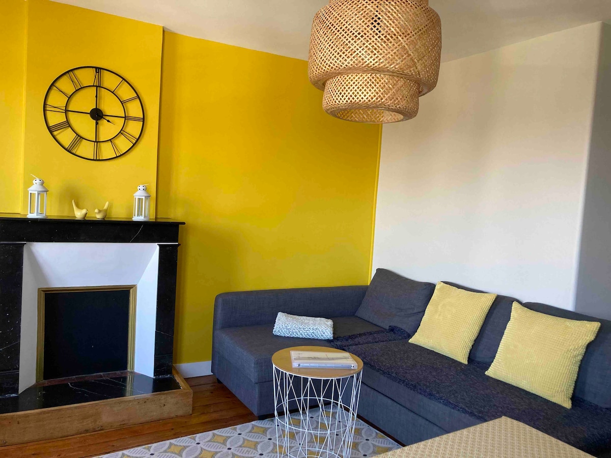 A bright living area features a large gray sectional sofa adorned with yellow cushions. A round coffee table rests on a light rug, and a modern clock is mounted on the vibrant yellow wall. A decorative fireplace adds a cozy touch to the space.