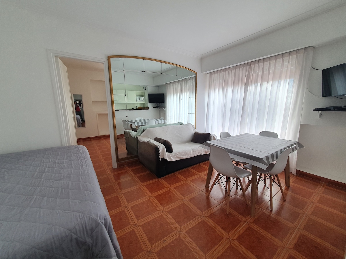 A bright living area features a spacious bed in the corner and a cozy sofa facing a dining table surrounded by chairs. Large windows with sheer curtains allow natural light to fill the space, while mirrored accents enhance the openness of the room.
