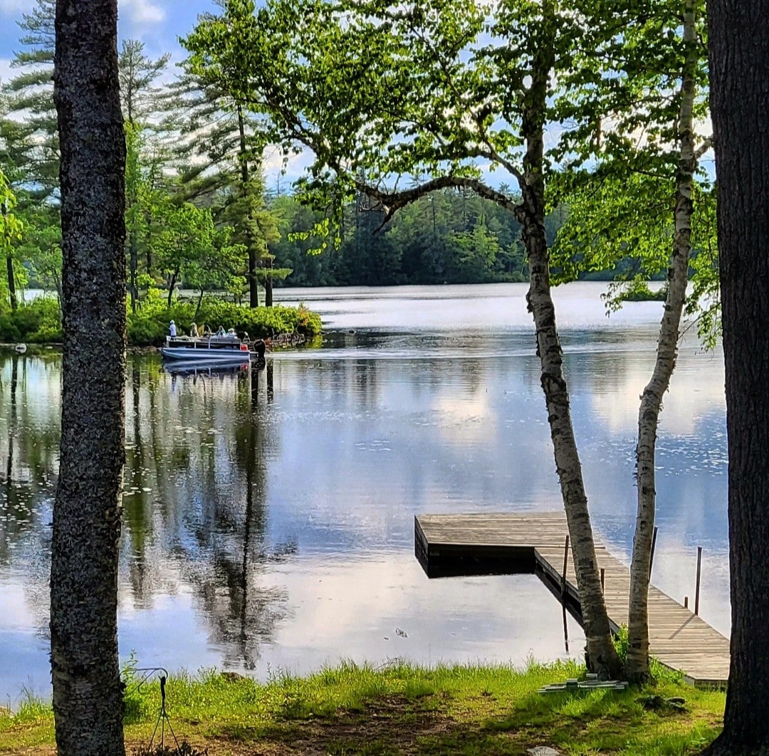 A serene view of Moose Pond is presented, featuring still waters reflecting surrounding trees and clouds. A private dock extends from the shore, with a boat visible alongside it. Lush greenery frames the scene, enhancing the tranquil lakeside atmosphere.