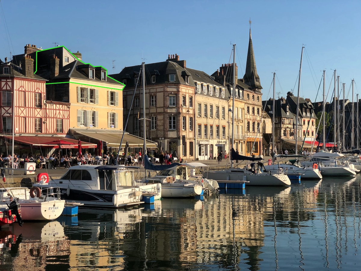 A charming row of historical buildings lines the waterfront, reflecting in the calm waters of the harbor. The scene features boats docked along the quay, with outdoor dining spaces visible, inviting guests to relax and enjoy the harbor view.