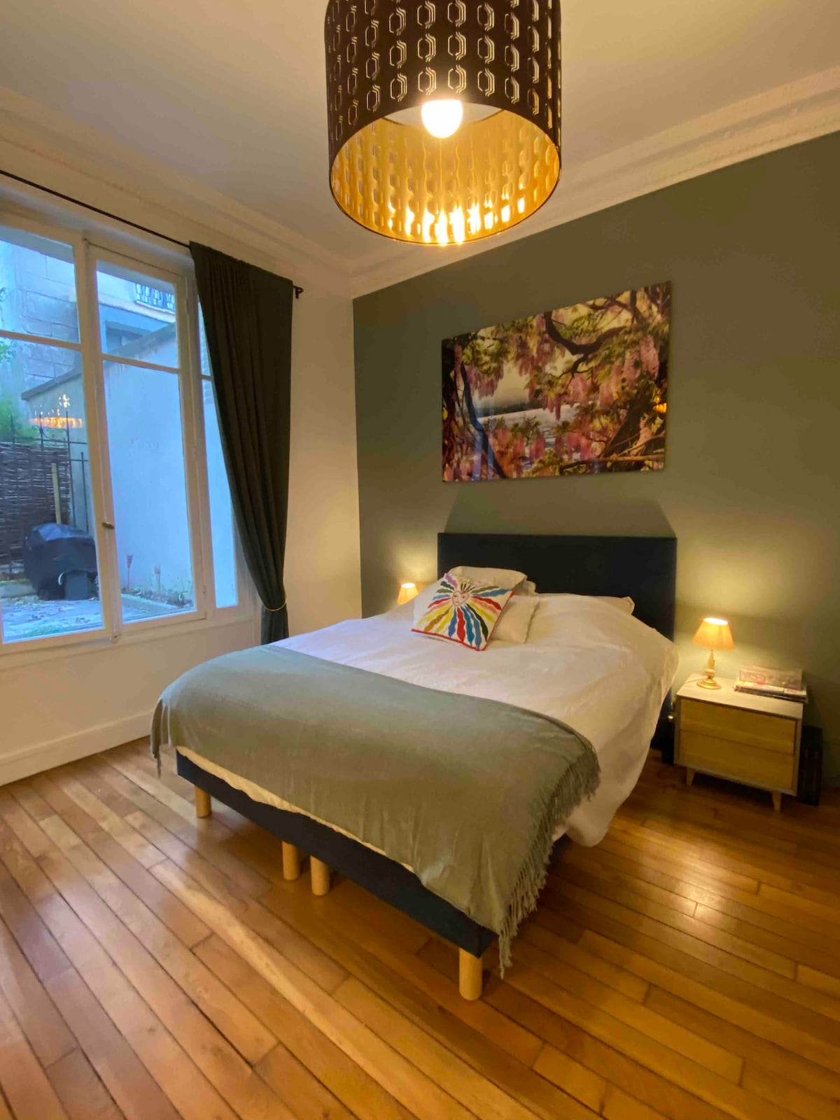 A sleeping area features a spacious bed against a green wall, complemented by a colorful wall art piece. Two bedside tables with lamps provide soft illumination. Natural light enters through a large window, and wooden flooring adds warmth to the room.