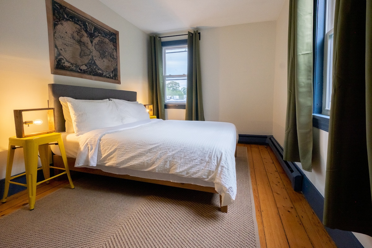 A private bedroom is presented with a spacious bed covered in white linens, complemented by a soft rug beneath. Natural light filters through two window panes, framed by green curtains, while a decorative wall art piece adds a touch of character. A yellow nightstand enhances the room's design.