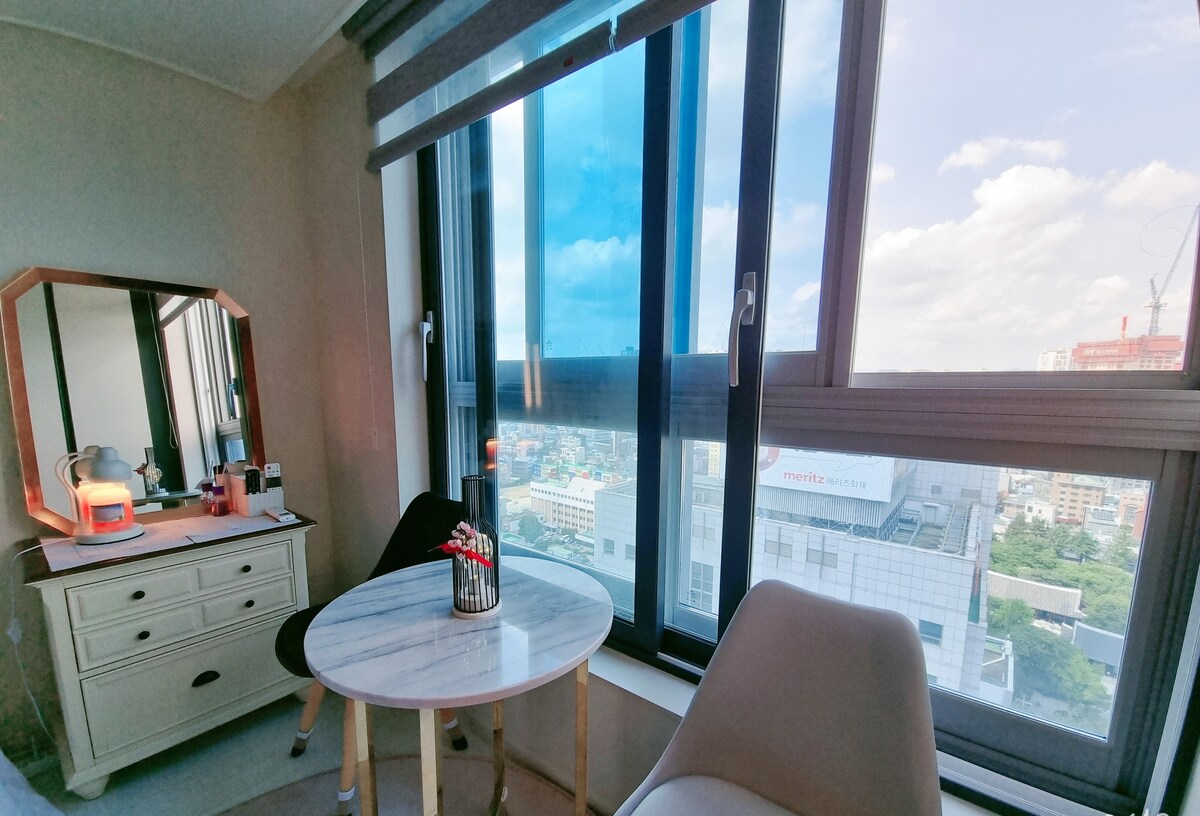 A bright corner of the room features large windows offering a view of the cityscape. A small round marble table is positioned between two chairs, with a lamp and various personal items on a nearby dresser. Natural light floods the space, creating an inviting atmosphere.