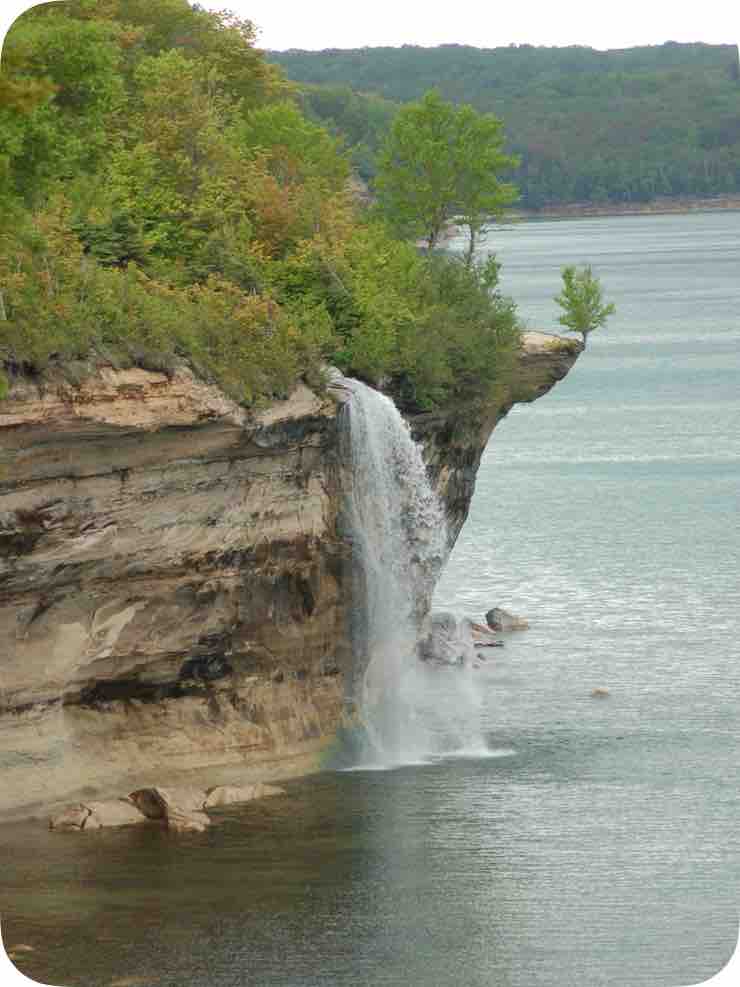 A waterfall cascades from a rocky cliff into a calm body of water, surrounded by lush greenery. The cliff exhibits various layers of textured rock, with trees peeking over the edge, showcasing nature's beauty in a serene setting.