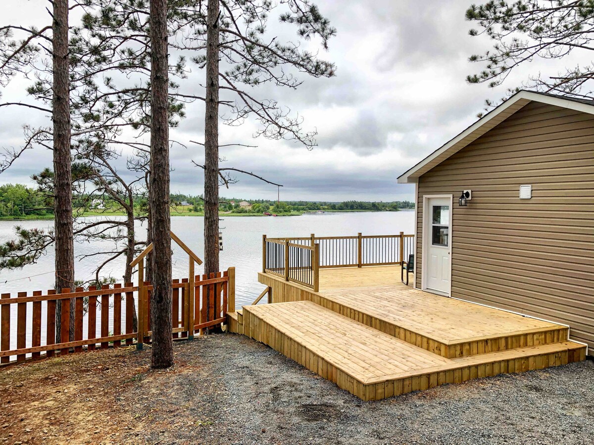 The exterior of the renovated chalet features a spacious deck made of wooden planks, with trees providing partial shade. The nearby water reflects a soft light, while the building displays a neutral-colored facade with a single door and window.