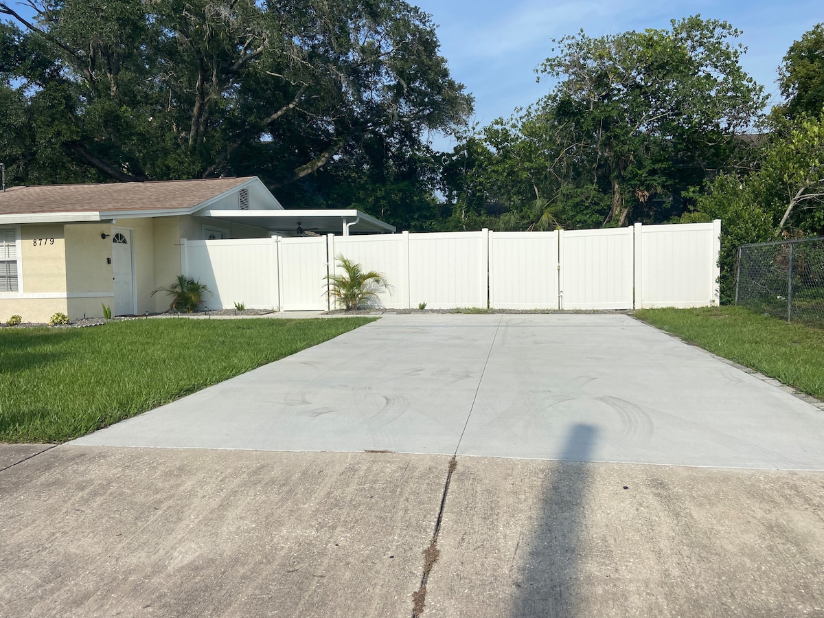 The image displays a spacious driveway with recently poured concrete, bordered by a neat green lawn. A white fence encloses the area, providing privacy. A single-story home is visible to the left, complemented by a well-kept entrance and minimal landscaping.