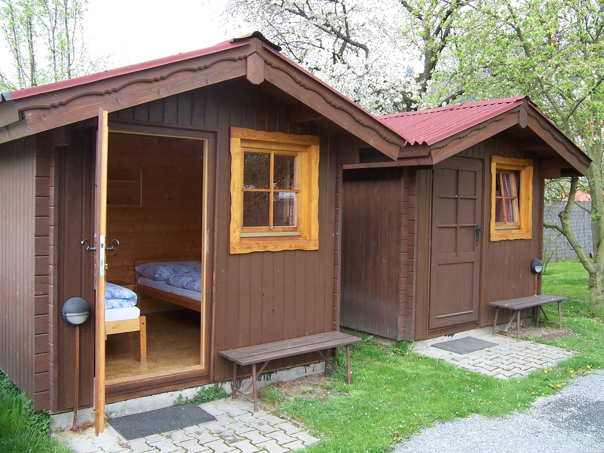 Two wooden bungalows are visible, each featuring a rustic design with sloped roofs and wooden accents. One bungalow has an open door, showcasing two beds inside. Benches are placed outside, and a path leads between the structures, surrounded by greenery and blossoming trees.