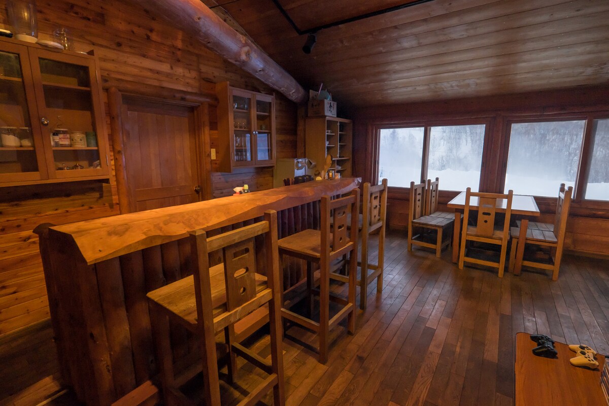 A wood-paneled common lounge space features a large bar made of polished wood and several wooden stools. Nearby, a dining table with chairs is positioned by expansive windows, allowing for views of the surrounding snowy landscape. The warm ambiance of the room is enhanced by wooden beams across the ceiling.