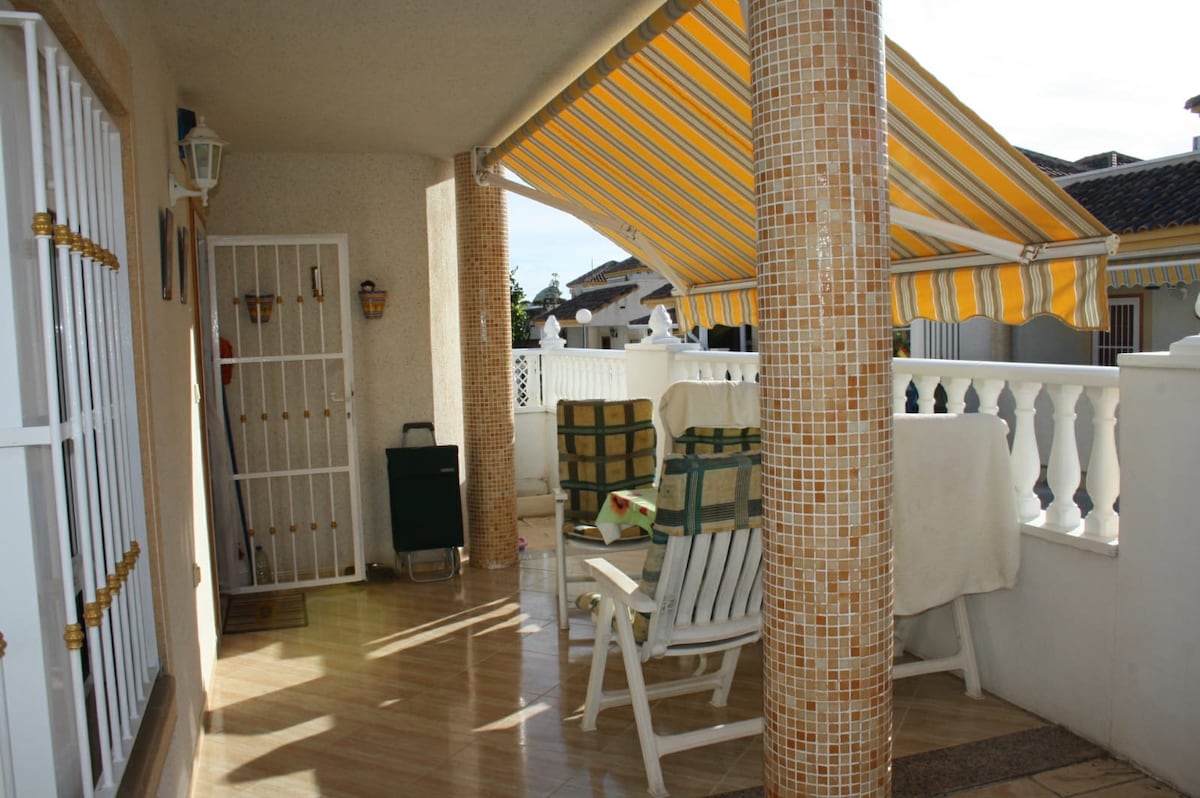 A covered terrace space is provided with a striped awning, featuring two lounge chairs and a small dining setup. The flooring is tiled, while the walls have decorative elements. Green plants and white railing enhance the outdoor ambiance.