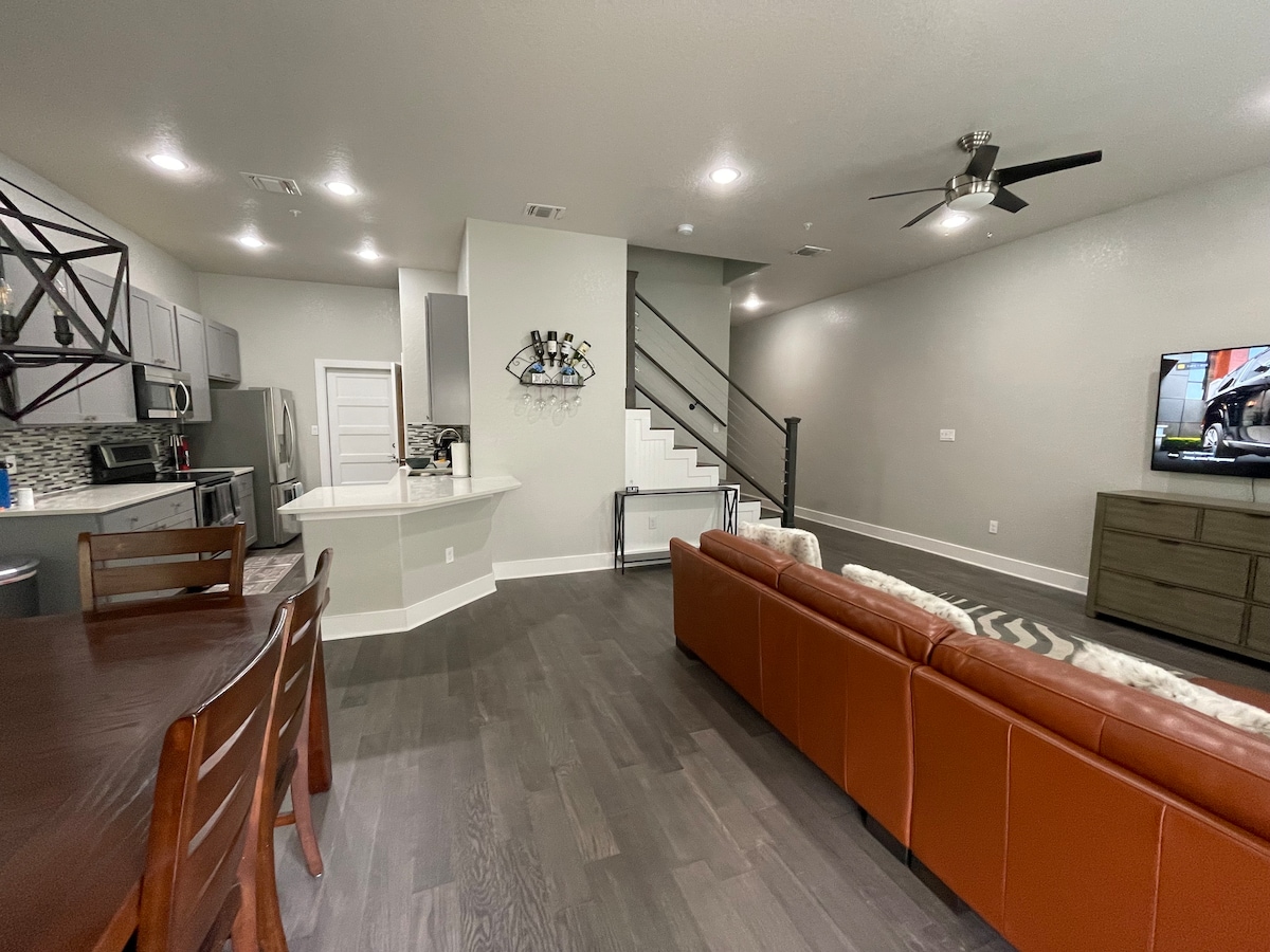 The living area features a spacious layout with a leather sofa facing a television. A dining table with several chairs is located nearby, while the kitchen includes modern appliances and a breakfast bar. A staircase leads to the upper level, enhancing the open design.