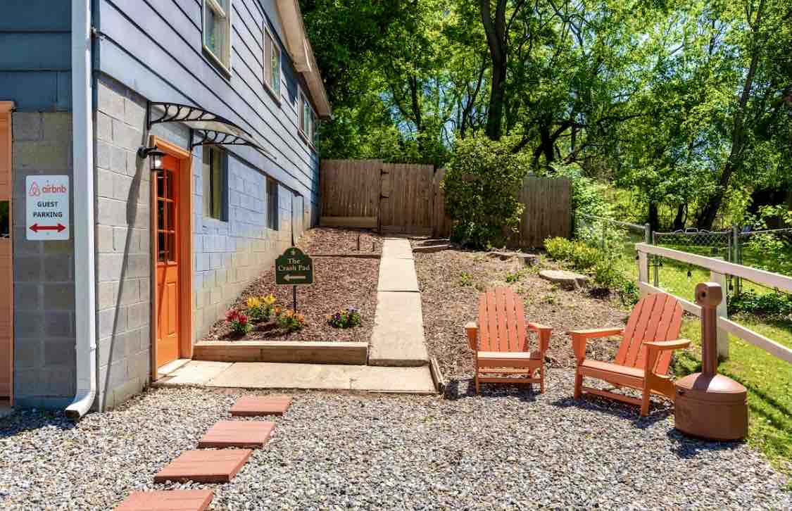 A private entrance is framed by vibrant orange doors, leading to a landscaped pathway. Two Adirondack chairs are positioned on gravel, inviting relaxation. Lush greenery and flowering plants enhance the outdoor setting, creating a serene ambiance in the South Slope neighborhood.