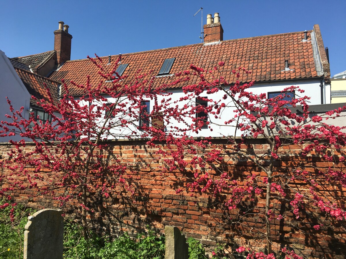 A vibrant wall adorned with flowering branches is showcased against a backdrop of traditional architecture. The brick wall displays a mix of textures, while windows with sloped roofs complement the scene under a clear blue sky.