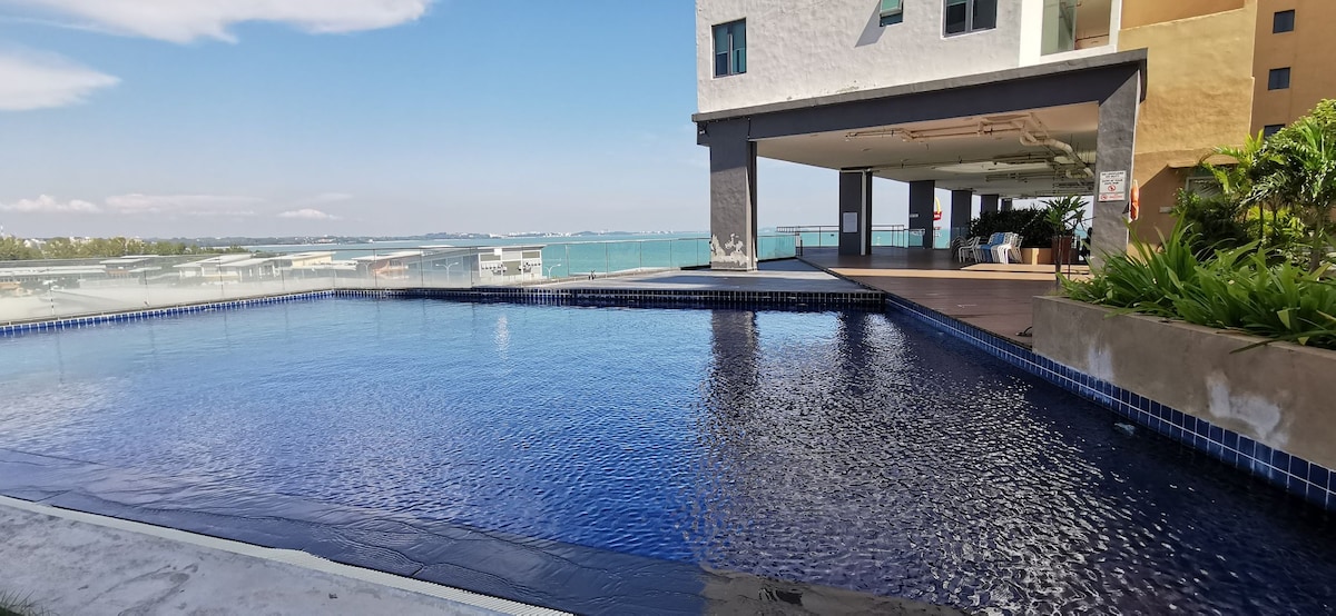 An outdoor infinity pool is presented, featuring calm water that reflects the sky. The pool is bordered by a stone deck and overlooks a scenic view of the sea and surrounding area. The structure above provides shaded seating space, enhancing the leisure experience.