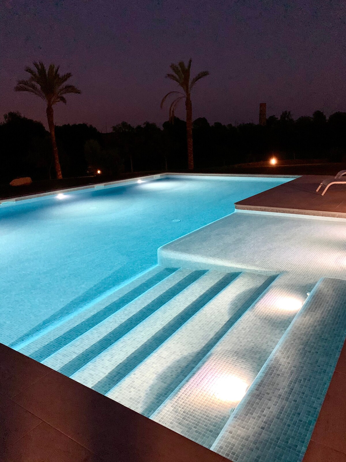 The illuminated pool glows in soft blue tones under the night sky, featuring cascading steps leading into the water. Palm trees are visible in the background, and ambient lighting enhances the tranquil atmosphere around the pool area.