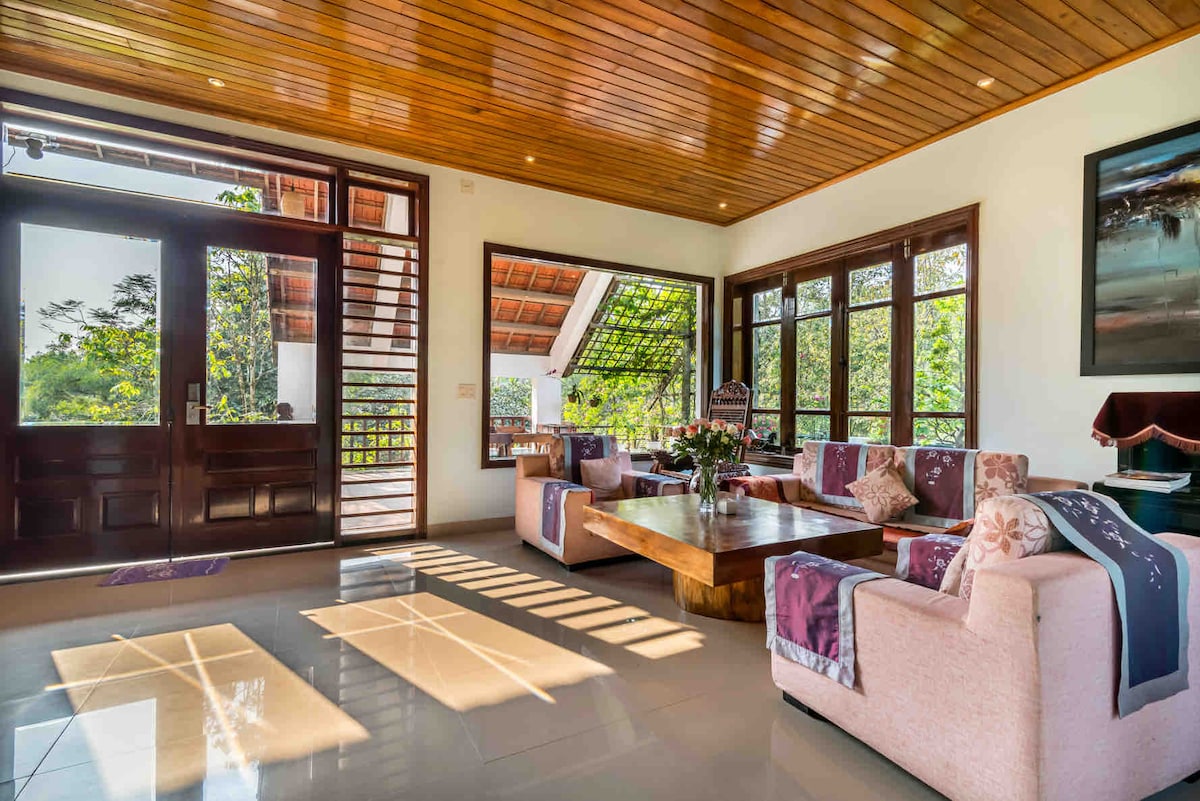 A well-lit living room features large windows inviting natural light. Comfortable seating is arranged around a wooden coffee table. A floral arrangement sits center stage, while the room opens to a view of greenery, enhancing the connection to the outdoors.