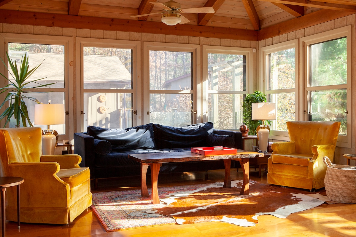A bright and airy living room features a dark blue couch, flanked by two vintage yellow armchairs. Natural light pours in through large windows, illuminating a rustic wooden coffee table and a vibrant area rug with a cowhide design.