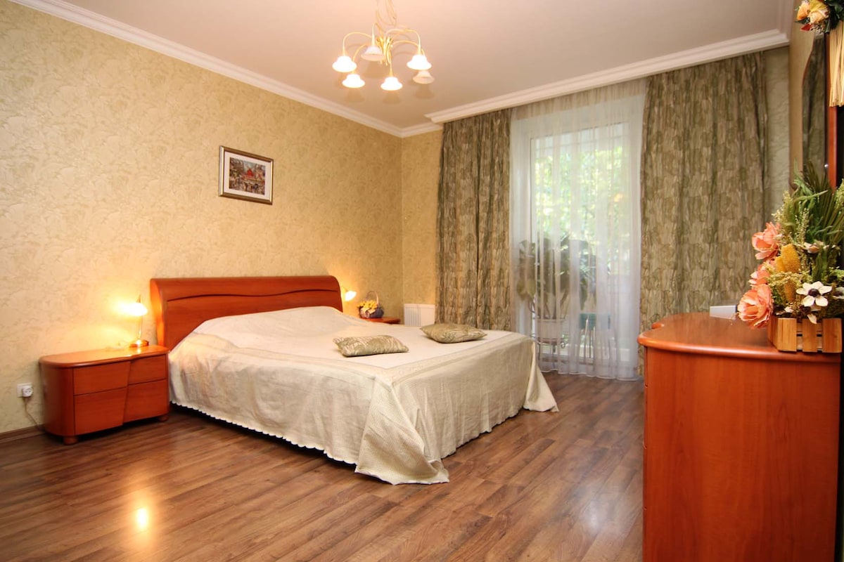 A spacious bedroom is depicted, featuring a large double bed covered with a light-colored blanket. Soft lighting is provided by a multi-bulb chandelier above. Large windows are adorned with sheer curtains, allowing natural light to fill the room, which also includes a wooden dresser and decorative plants.