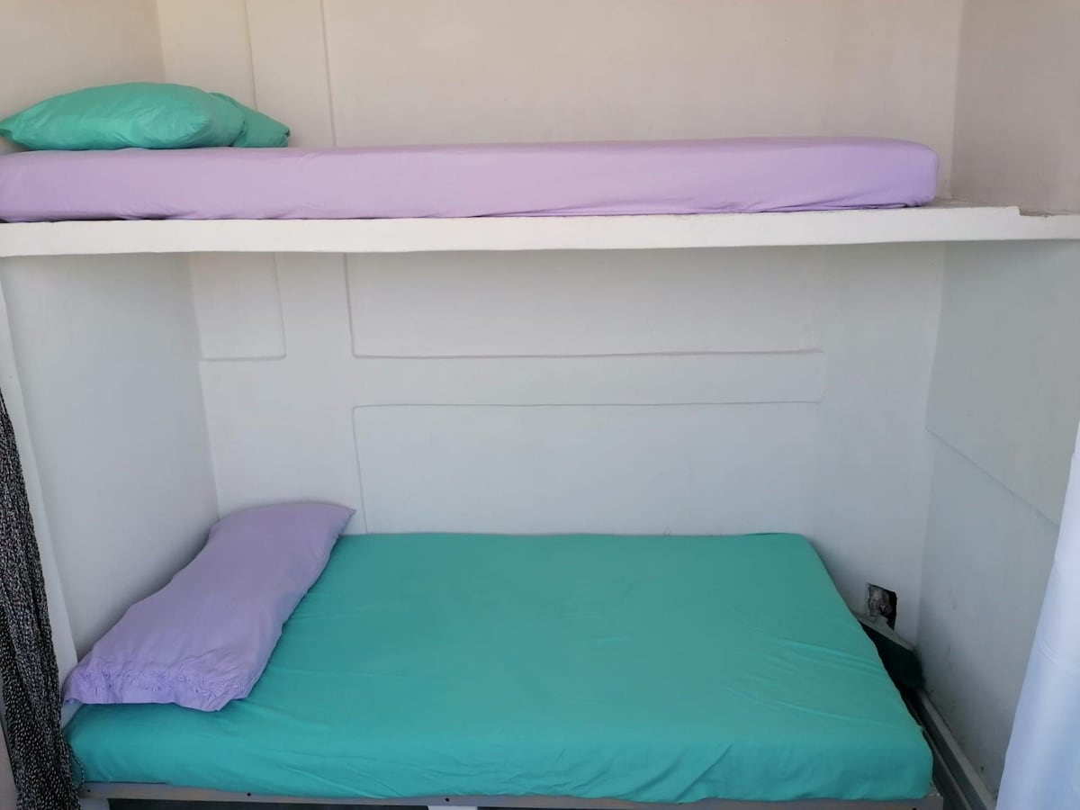 A compact sleeping area is arranged with two elevated beds, each featuring a light purple and turquoise bedding set. Shelving above and below provides additional space, while the minimalist walls enhance the room's simplicity.