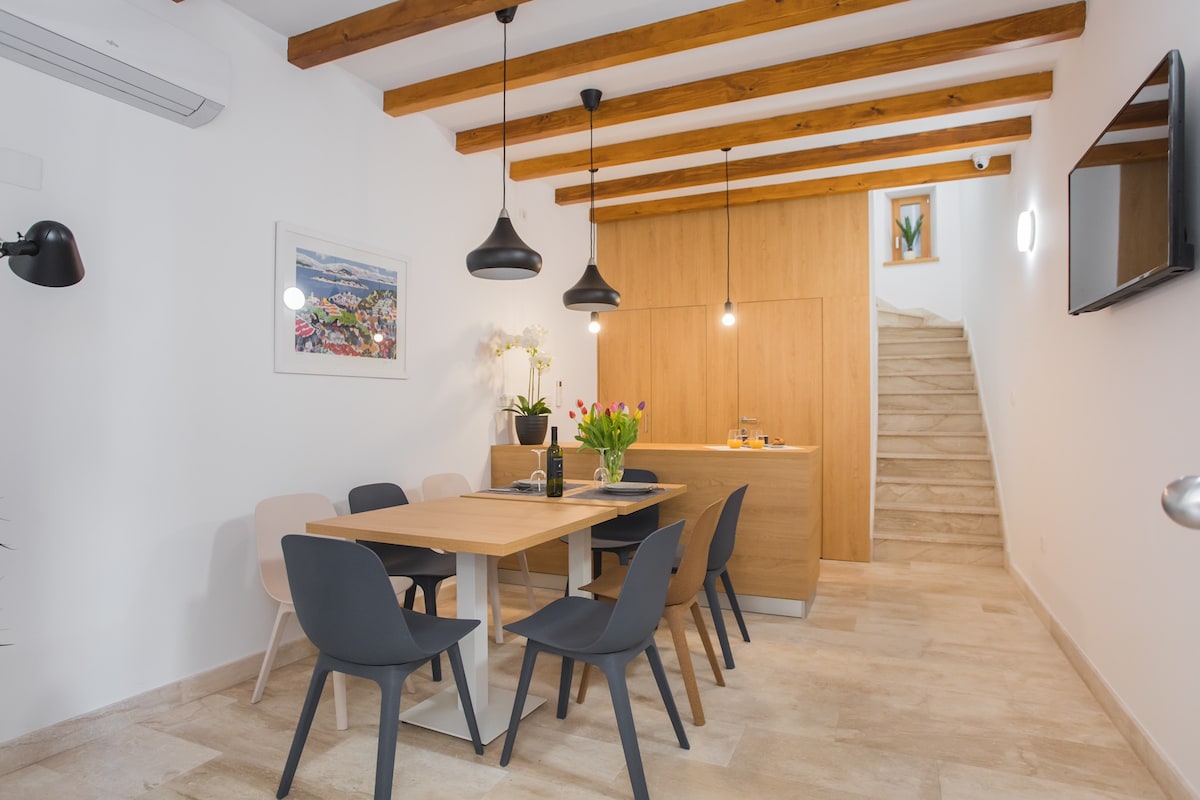 A contemporary dining area is presented, featuring a wooden table surrounded by six chairs. Above, modern pendant lights hang from the exposed wooden beams. A kitchenette is visible in the background, along with stairs leading to an upper level. Fresh flowers sit at the center of the table.