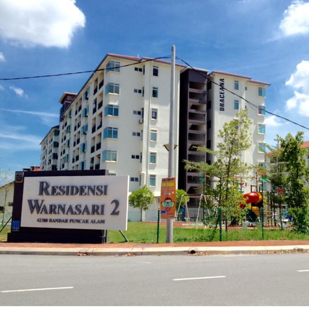 The exterior of a multi-story residential building is visible, featuring a sign for 'Residensi Warnasari 2.' The structure is framed by a clear blue sky, with a playground area nearby, showcasing various colorful play equipment set against a backdrop of greenery.