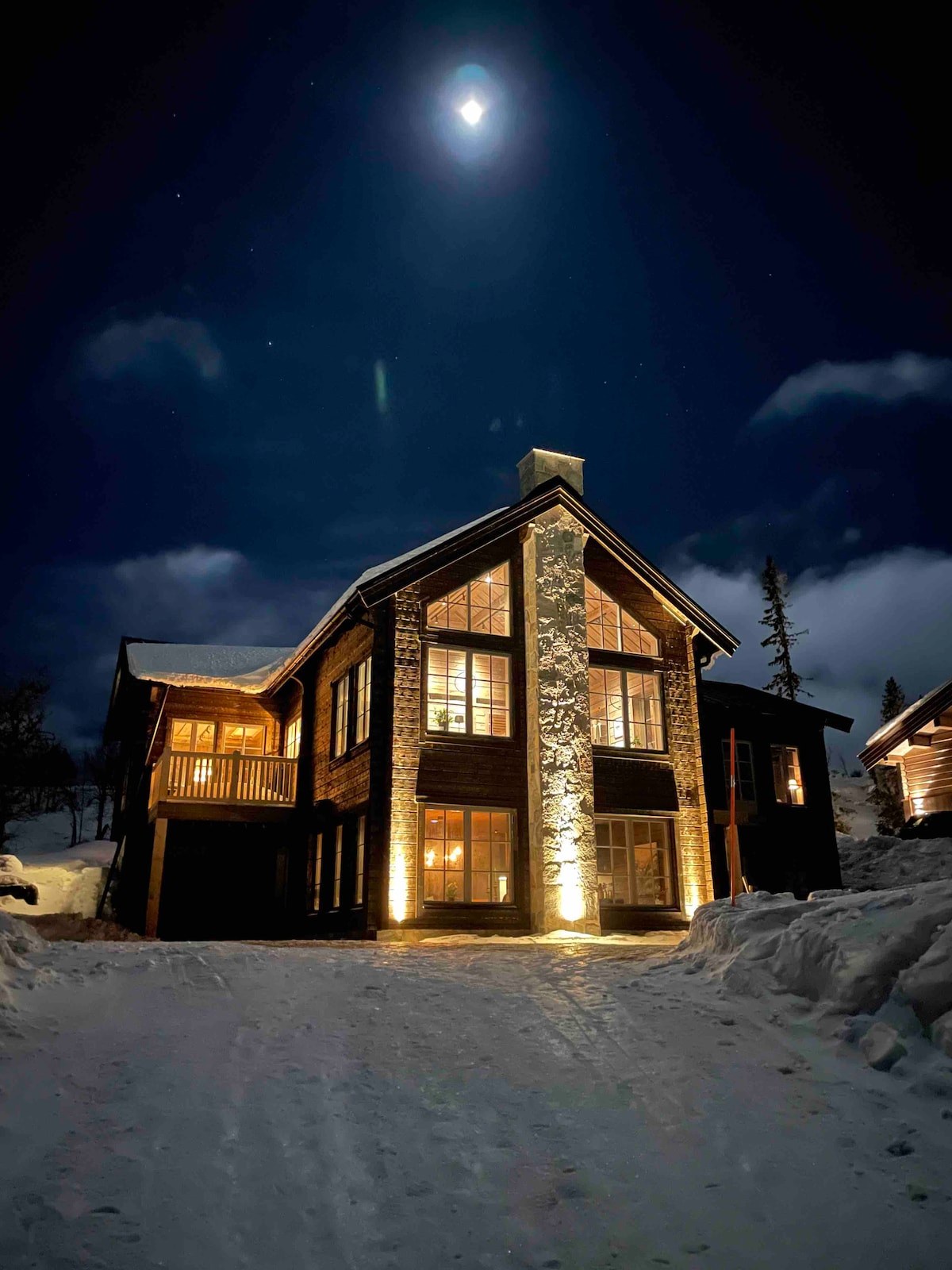 The exterior of the lodge is illuminated under a night sky, highlighting the wooden structure and stone accents. Snow blankets the ground, creating a serene atmosphere. The large windows reflect warm light, suggesting inviting spaces inside.