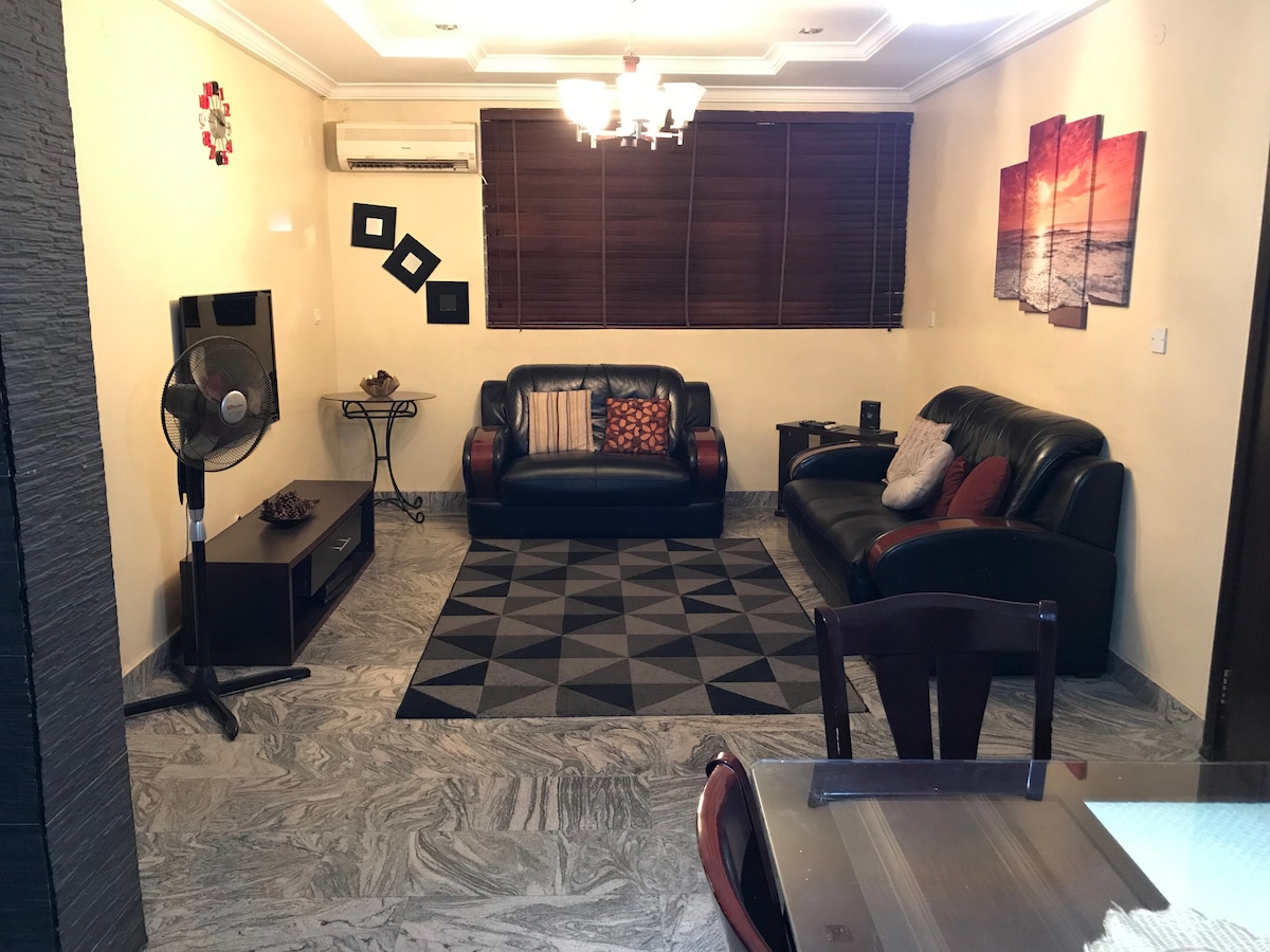 The living area features two leather sofas positioned around a geometric patterned rug. A small table with decorative items and a wall-mounted television are present. Wooden blinds filter natural light, while a ceiling light fixture provides illumination. A fan is placed in the corner.