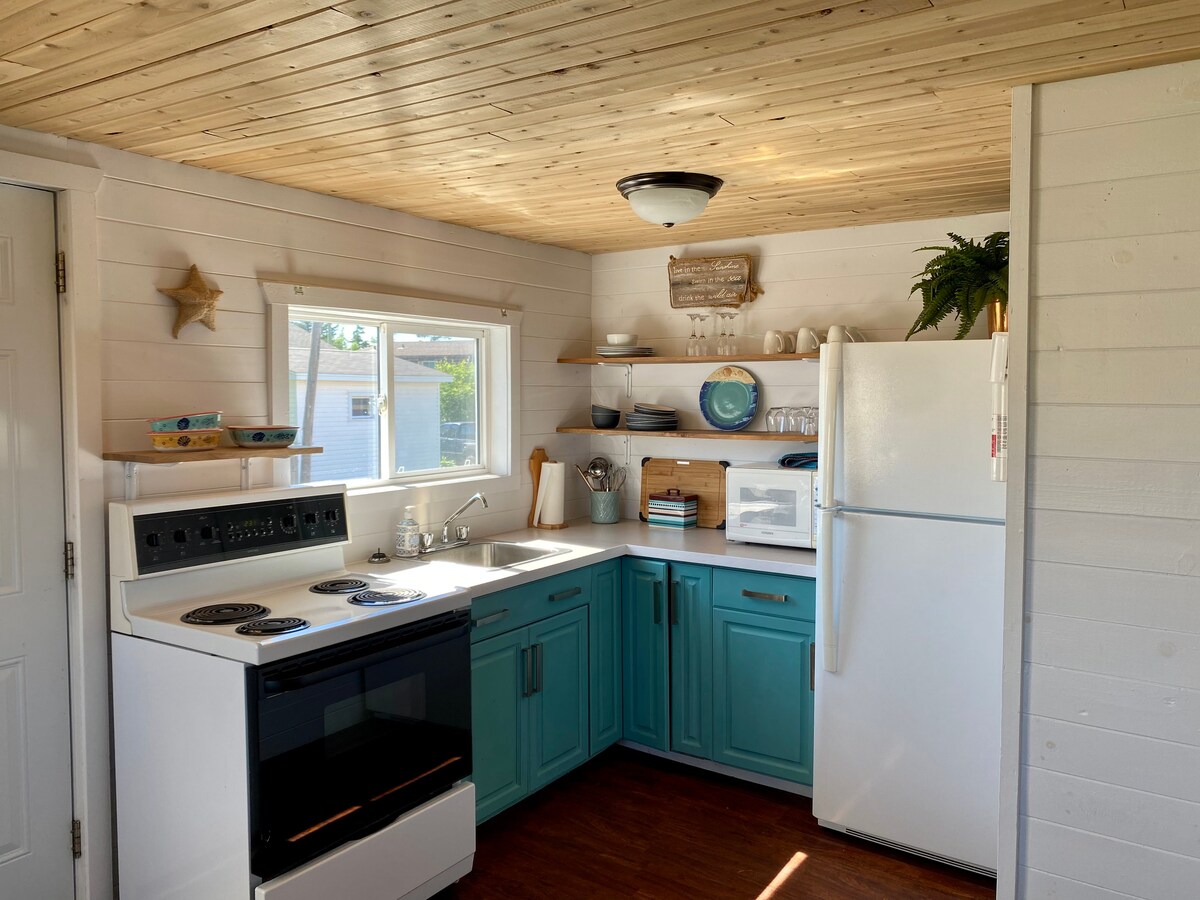 A bright kitchen features white cabinets with turquoise accents. An electric stove and a refrigerator are prominently displayed. Open shelving showcases dishware and decorative items. A window allows natural light to fill the space, enhancing the clean and functional design.