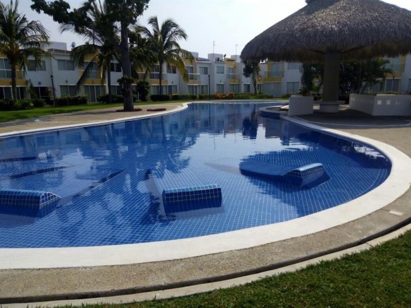 A tranquil swimming pool is surrounded by lush green grass and palm trees. Lounge chairs are set within the water, providing a relaxing spot. A thatched-roof gazebo is visible nearby, offering shade and leisure space. The area is well-maintained, encouraging a peaceful atmosphere.