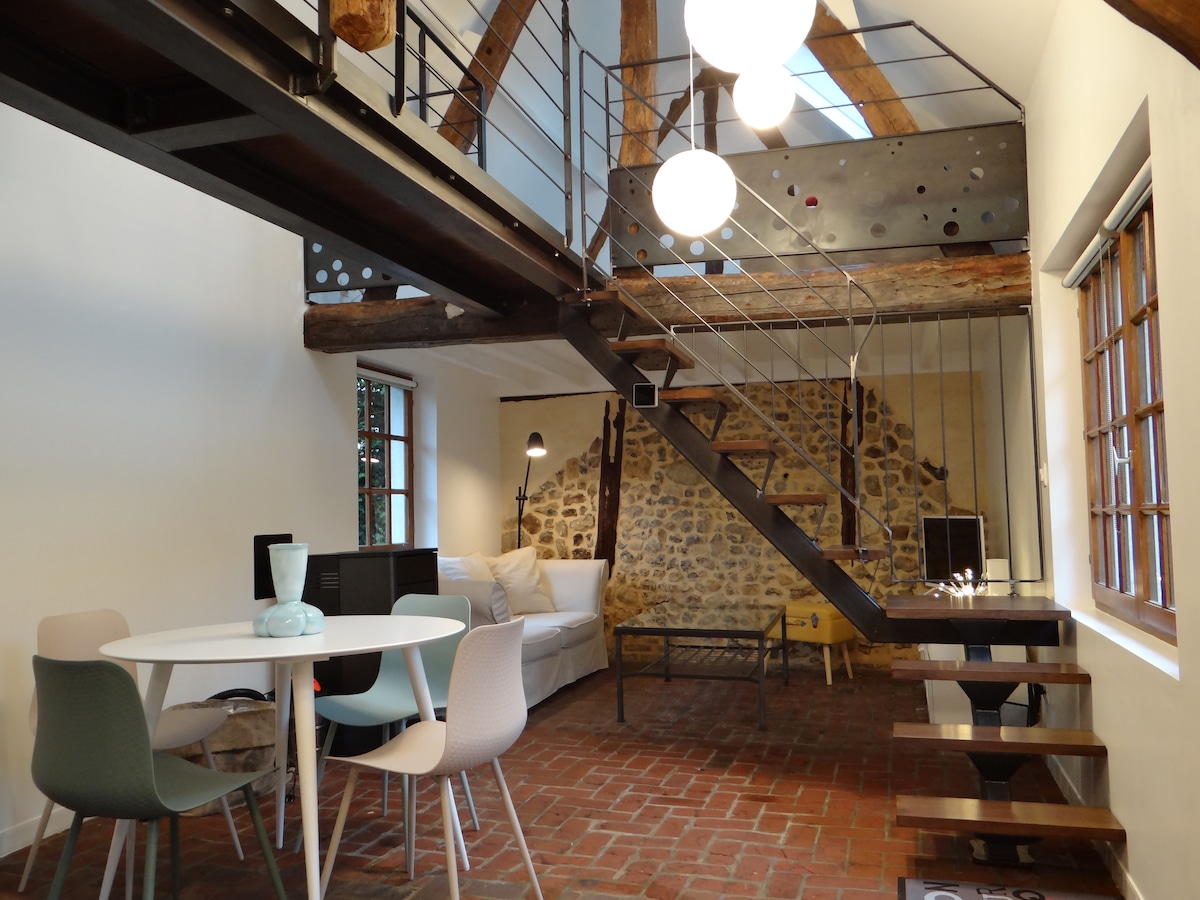 An open living space features a cozy seating area with a light sofa and a wooden coffee table. Modern dining chairs surround a round table. Exposed wooden beams accent the ceiling, while a staircase leads to a mezzanine level, enhancing the room's spaciousness.