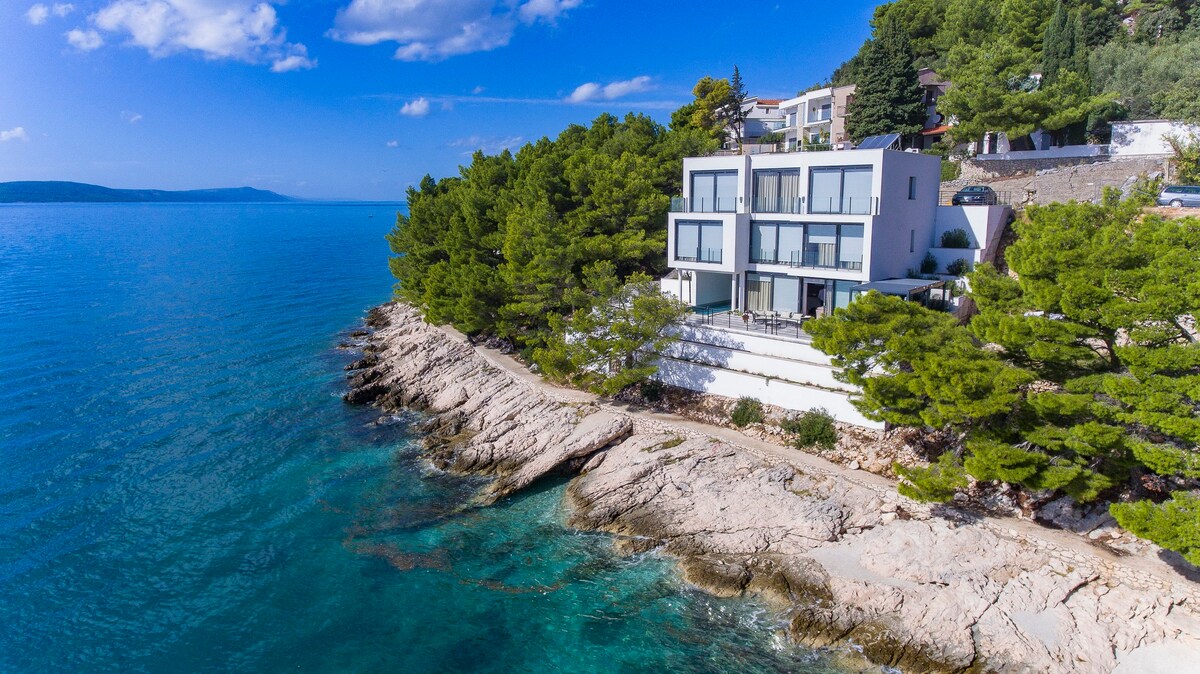 A modern villa is situated on a rocky coastline, surrounded by lush greenery. The structure features large windows, offering views of the serene sea. The clear blue water gently laps against the shore, creating a peaceful coastal ambiance.