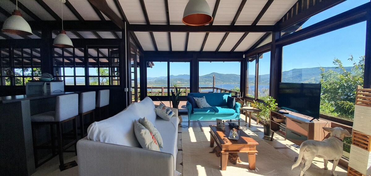 A bright and airy living area features large glass windows that provide views of the surrounding landscape. Comfortable seating includes a light-colored sofa and a teal blue couch. A wooden coffee table is centered on a light rug. A television is positioned within the space.