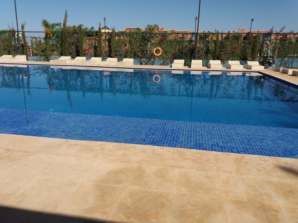 A clear blue swimming pool is set against a backdrop of lush greenery. Sun loungers line the pool's edge, providing ample space for relaxation. The surrounding area is well maintained, with decorative elements enhancing the tranquil atmosphere.