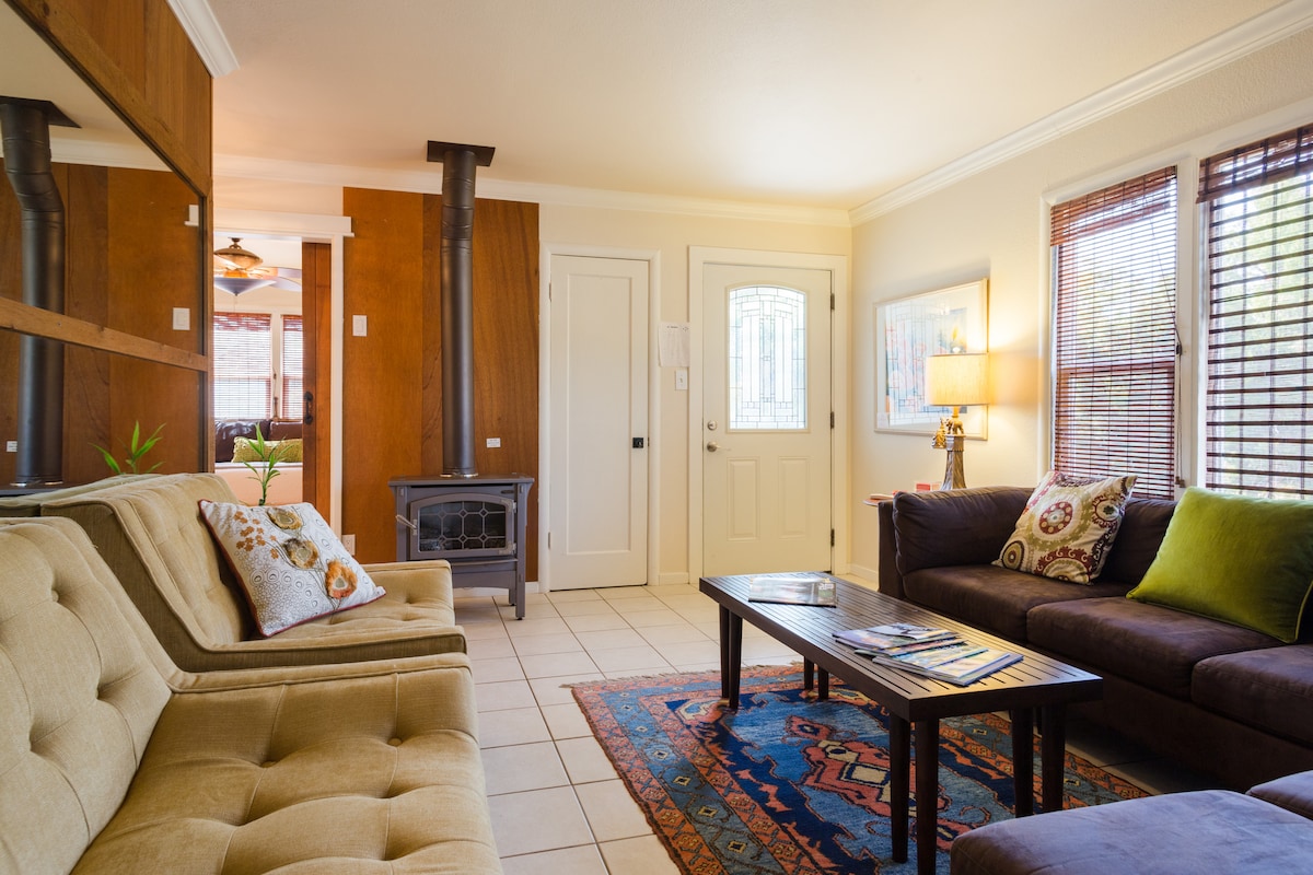 The living room features a cozy seating arrangement with a light-colored sofa and a dark sectional couch. A coffee table sits in the center on a patterned area rug. Natural light fills the space through windows, with a decorative wall mirror and a wood-burning stove adding warmth.