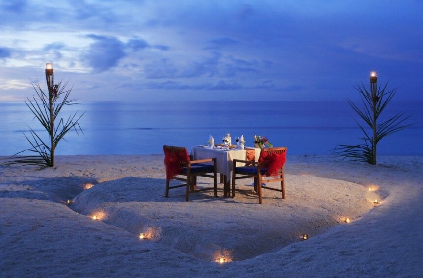 A beach dining setup is presented on a sandy area, illuminated by soft candlelight. A round table is set for two, surrounded by colorful chairs. Palm fronds are positioned on either side, with a serene ocean view fading into a twilight sky.
