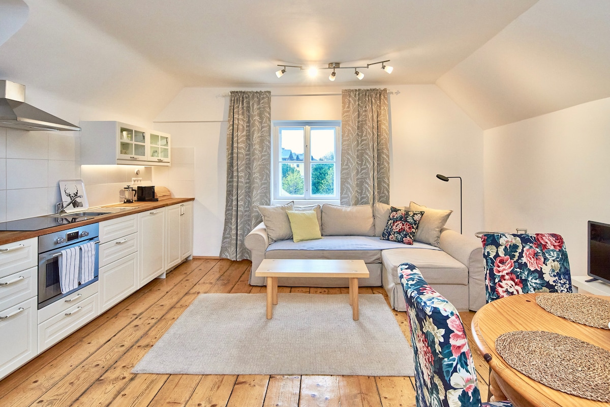 A bright living area features a comfortable sofa adorned with floral patterns and a coffee table in the center. Natural light pours in through a window dressed with light curtains, illuminating the wooden flooring and the adjacent dining table with chairs.