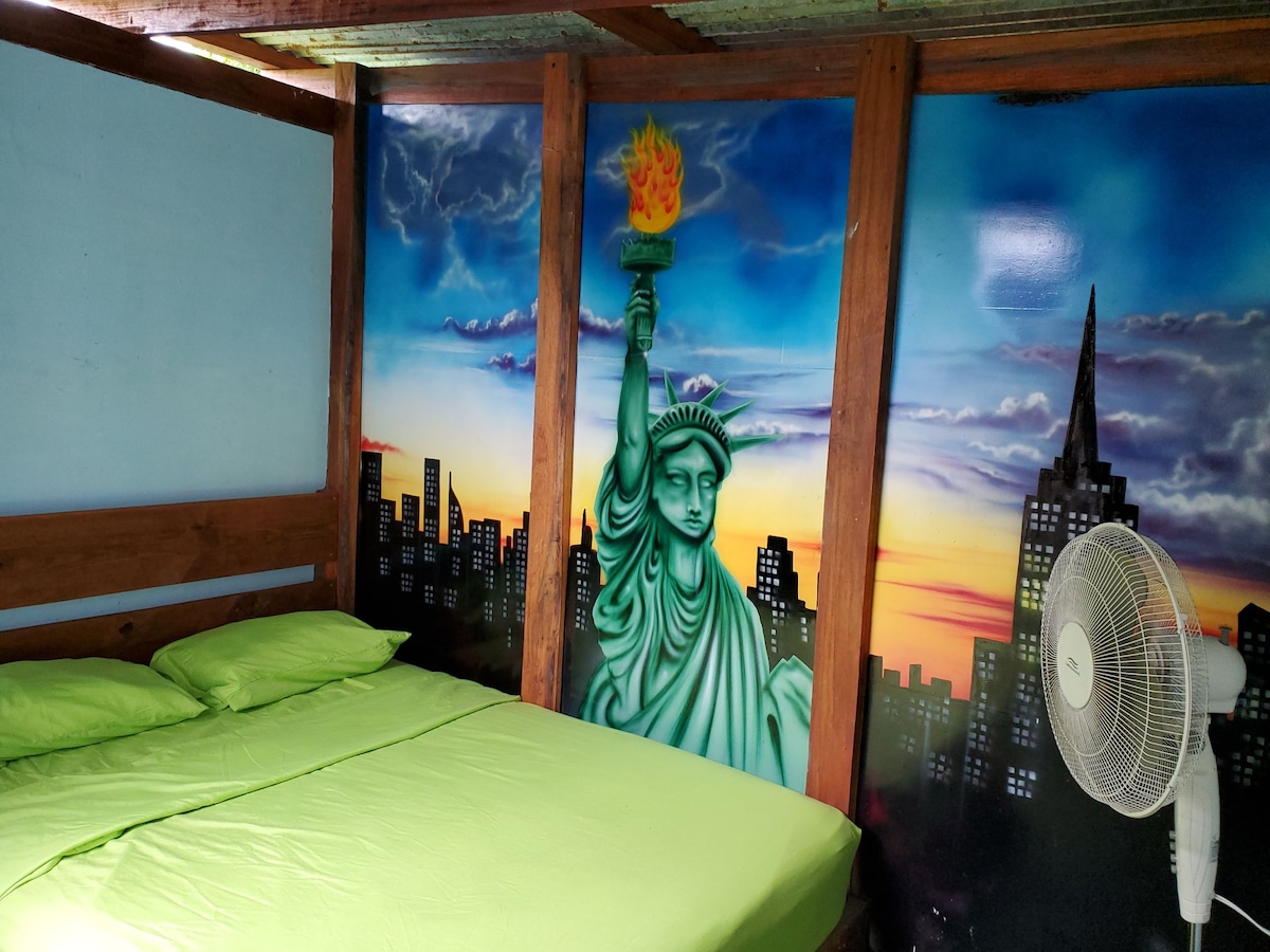 A sleeping area features a spacious bed covered with bright green sheets. A mural depicting the Statue of Liberty stands on one wall, complemented by a vibrant city skyline and a colorful sunset. A ceiling fan is positioned nearby, providing ventilation.