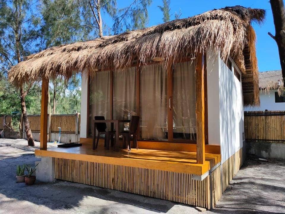A charming bungalow features a thatched roof and a spacious wooden deck. Large glass windows allow natural light to fill the interior. A small table and chairs are placed outside, inviting relaxation in a tranquil outdoor setting surrounded by greenery.