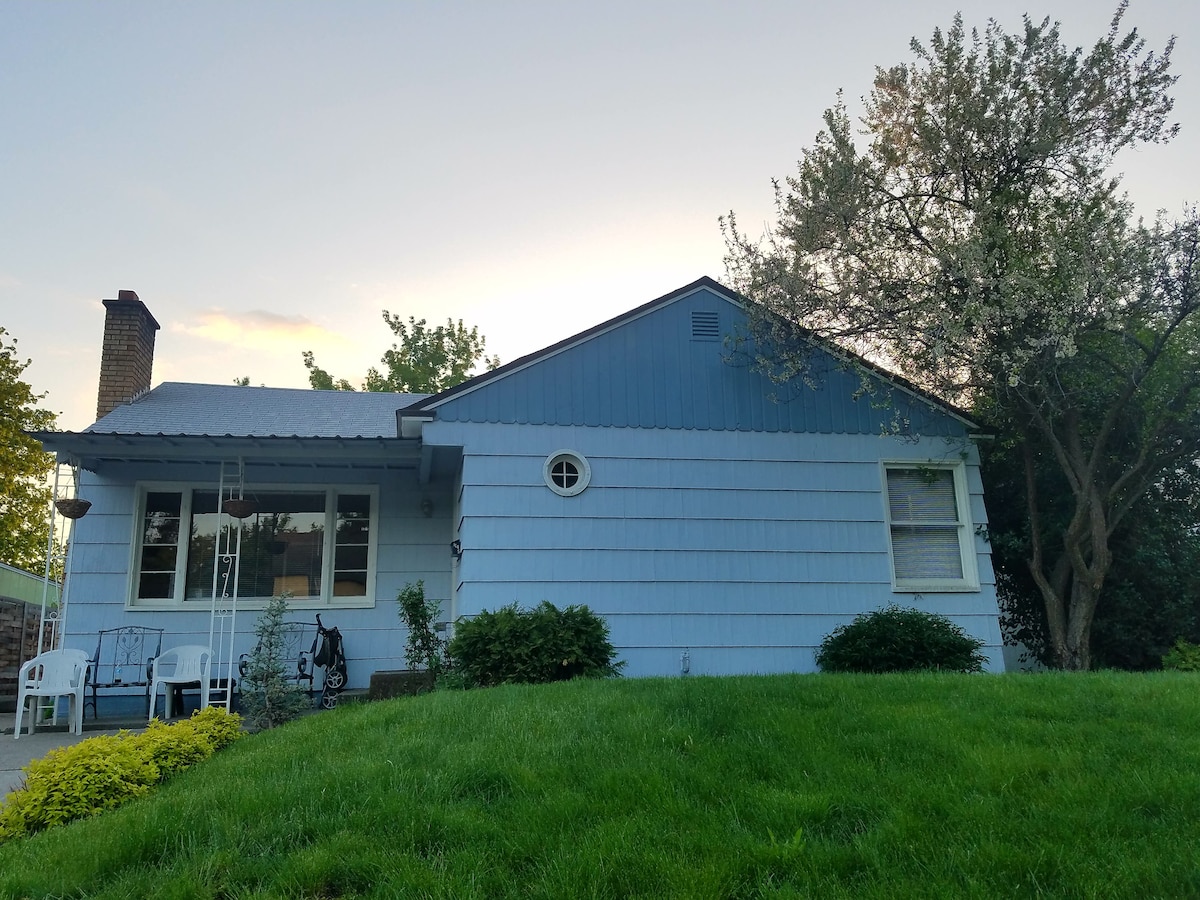 The exterior of the bungalow showcases a light blue facade beneath a soft sunset sky. A well-maintained lawn frames the house, complemented by lush green bushes and a flower bed. A cozy seating area with white chairs is visible on the porch.