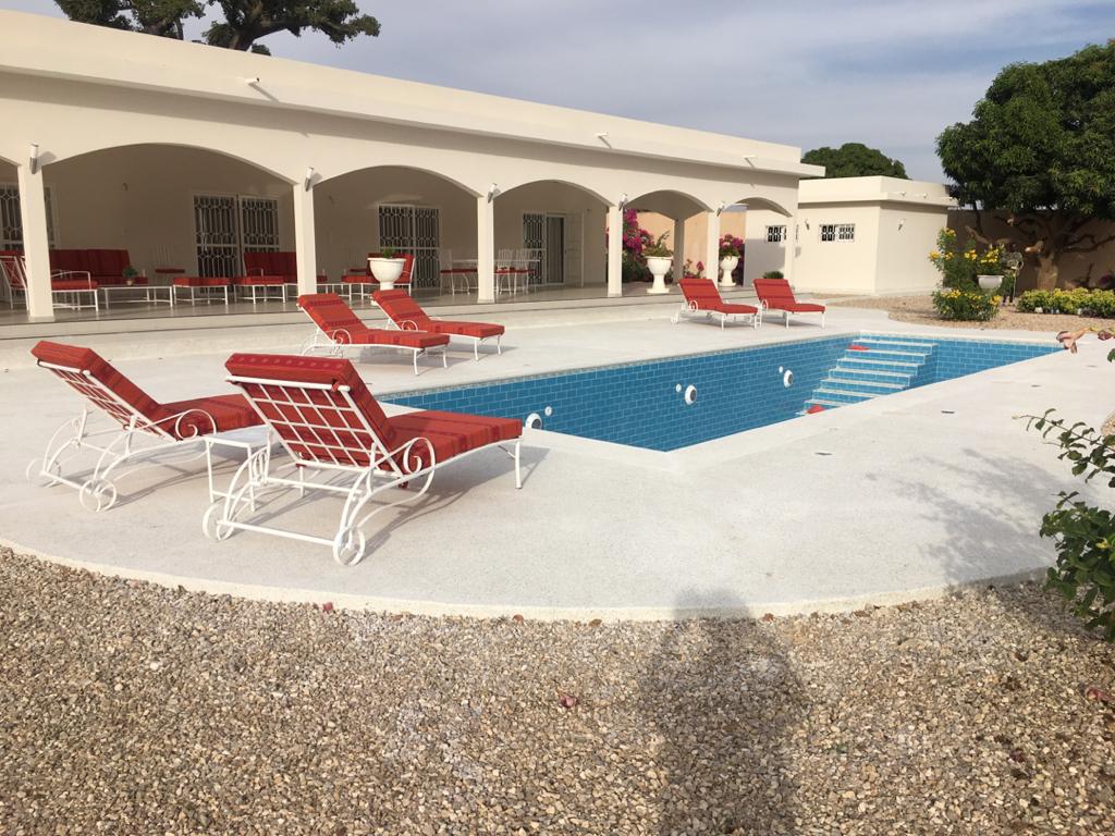 A solar-heated swimming pool is surrounded by a spacious deck featuring several lounge chairs. The pool area is bordered by landscaped gardens and an expansive terrace with shaded seating. Arched openings lead to the interior of the property, providing views of vibrant flora.