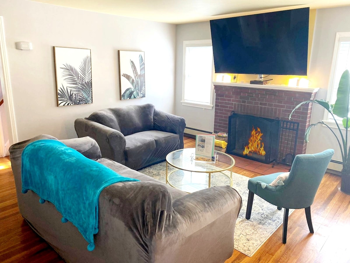A spacious living room features a large flat-screen TV mounted above a classic fireplace. Comfortable seating is provided by a sofa, loveseat, and accent chair. Two windows allow natural light to fill the space, complemented by decorative wall art and a round glass coffee table.