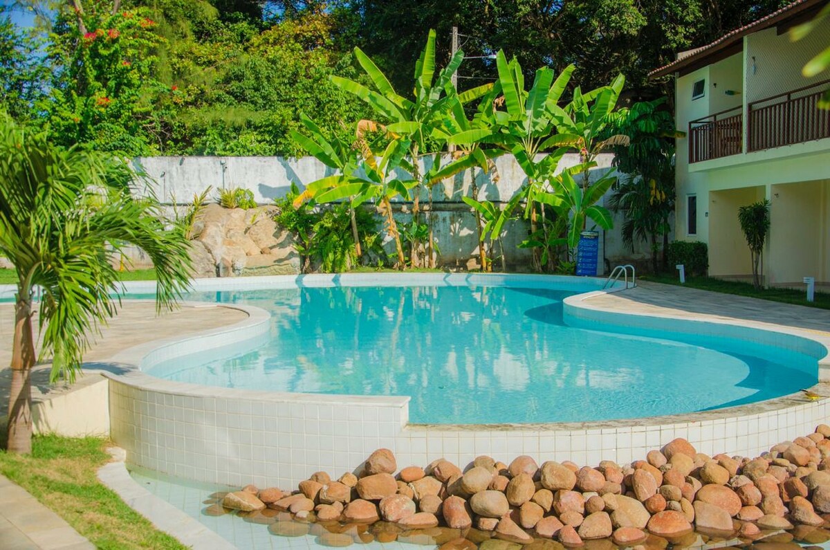 A serene pool is surrounded by tropical vegetation, featuring smooth stones lining its edge. The clear water reflects sunlight, creating a tranquil atmosphere. Lush banana plants provide shade, enhancing the outdoor space for relaxation and leisure.