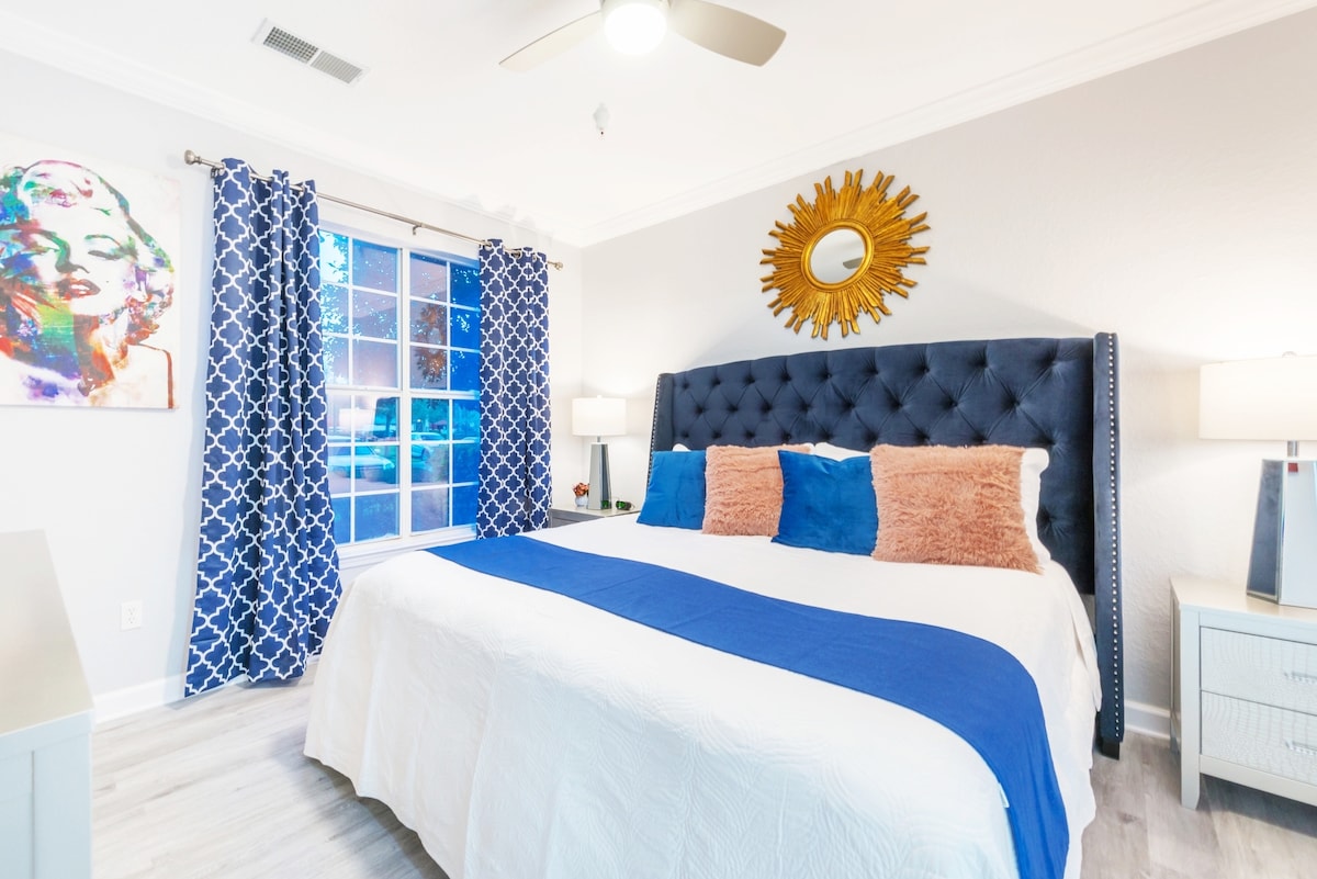 A contemporary bedroom features a spacious bed with a blue throw blanket and decorative pillows. A tufted blue headboard adds style, while sunlight streams through a large window adorned with patterned curtains. A circular mirror and bedside lamps enhance the modern decor.