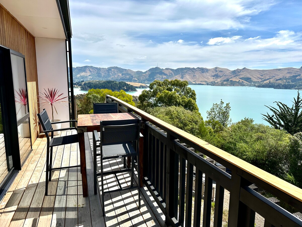 A spacious deck is shown, furnished with a wooden table and four chairs. A serene view of the lake and surrounding hills is visible in the background, with clear skies and gentle greenery framing the scene.