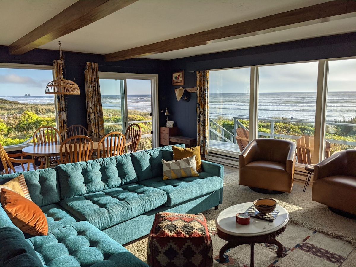 A cozy living area showcases a large teal sectional sofa adorned with accent pillows. Floor-to-ceiling windows present unobstructed ocean views, while the adjacent dining area features a wooden table surrounded by matching chairs. Natural light creates a bright and inviting space.